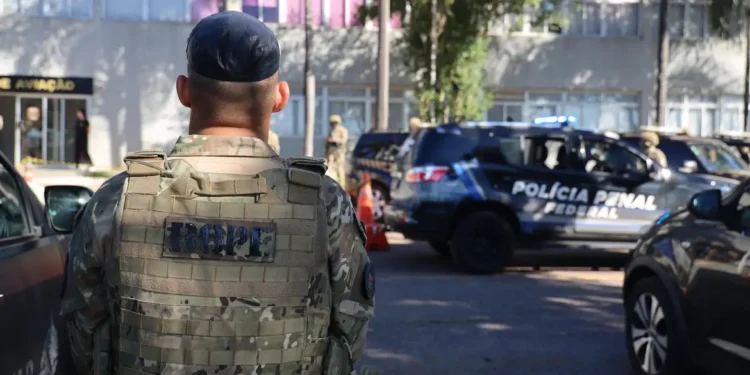 Agente da tropa com uniforme camuflado de costas próximo a viaturas da Polícia Penal Federal em operação