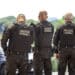 Três agentes da polícia penal vistos de costas com coletes táticos e armas em operação externa