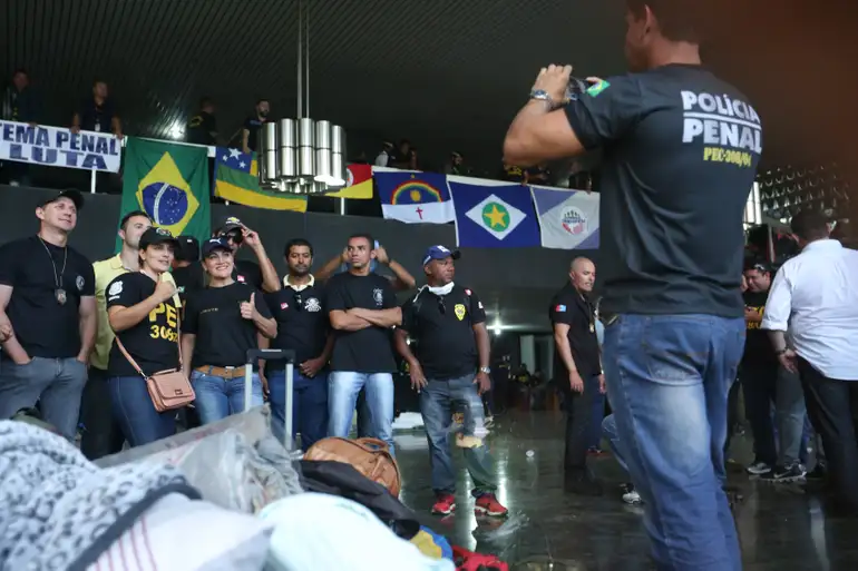 Grupo de policiais penais reunidos em protesto com bandeiras e cartazes em ambiente fechado