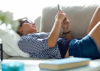 Pessoa relaxando no sofá e usando o celular, ilustrando a procrastinação.