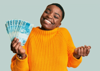 Mulher sorridente vestindo suéter amarelo segurando várias notas de 100 reais.