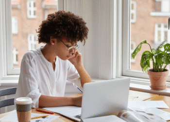 Mulher concentrada falando ao telefone e fazendo anotações com laptop aberto perto da janela.