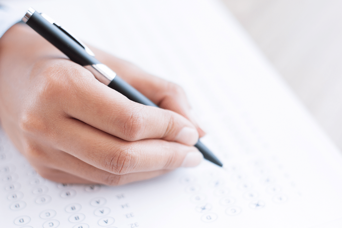 Mão segurando caneta preta preenchendo respostas em gabarito de prova objetiva com bolinhas