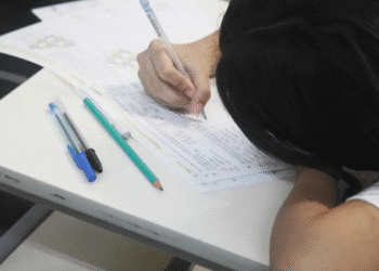 Estudante com cabelo escuro escrevendo em prova sobre mesa branca com canetas ao lado