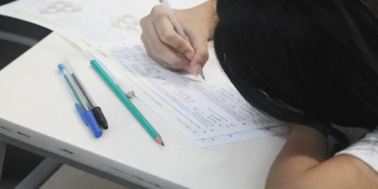 Estudante com cabelo escuro escrevendo em prova sobre mesa branca com canetas ao lado