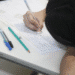 Estudante com cabelo escuro escrevendo em prova sobre mesa branca com canetas ao lado