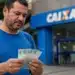 Homem confere cédulas de real em frente a uma agência da Caixa, representando a solicitação do saque do FGTS até 22 de julho.