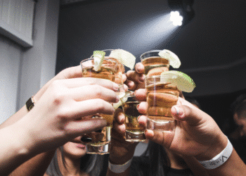 Mãos segurando copos de shot com bebida dourada e fatias de limão em ambiente de festa
