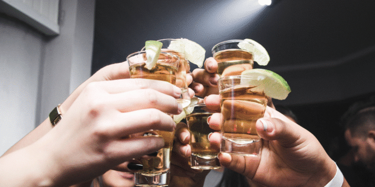Mãos segurando copos de shot com bebida dourada e fatias de limão em ambiente de festa