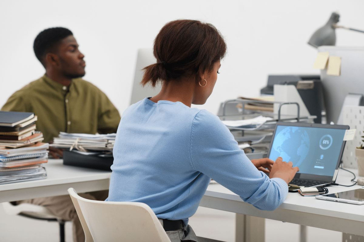 Duas pessoas sentadas em escritório, uma mulher usando laptop com gráficos na tela, homem ao fundo com pilha de documentos
