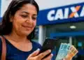 Mulher consulta o celular enquanto segura notas de dinheiro em frente a uma agência da Caixa, representando as novas vagas do auxílio Mães de Pernambuco em 2026.