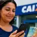 Mulher consulta o celular enquanto segura notas de dinheiro em frente a uma agência da Caixa, representando as novas vagas do auxílio Mães de Pernambuco em 2026.