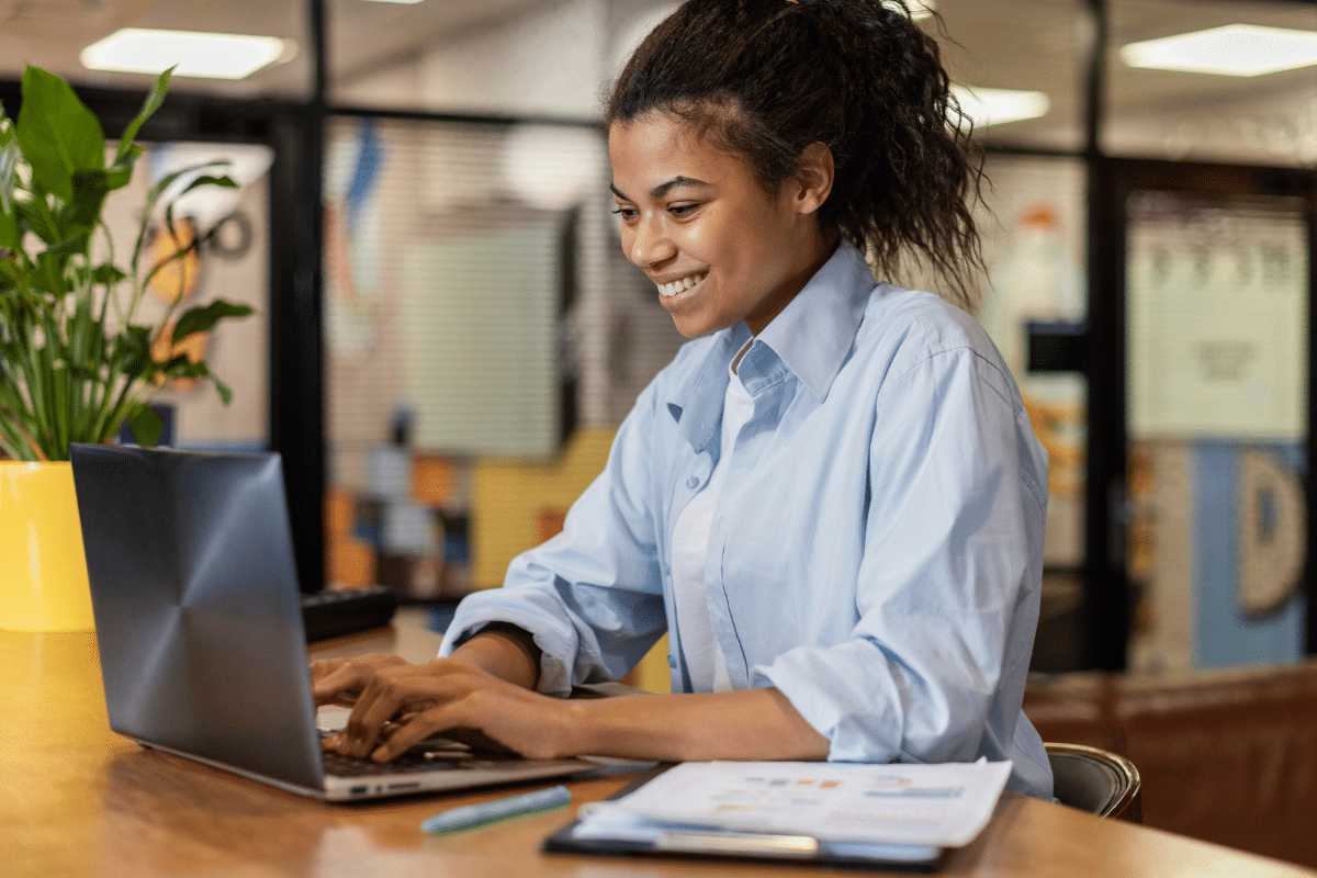 Mulher jovem sorrindo enquanto digita em laptop em mesa de trabalho com documentos e planta ao lado