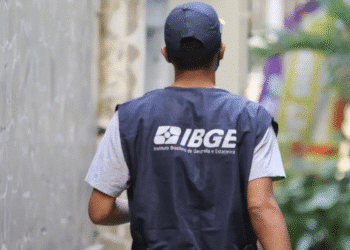 Agente do IBGE de costas com colete azul escuro e boné, caminhando entre prédios para coleta de dados