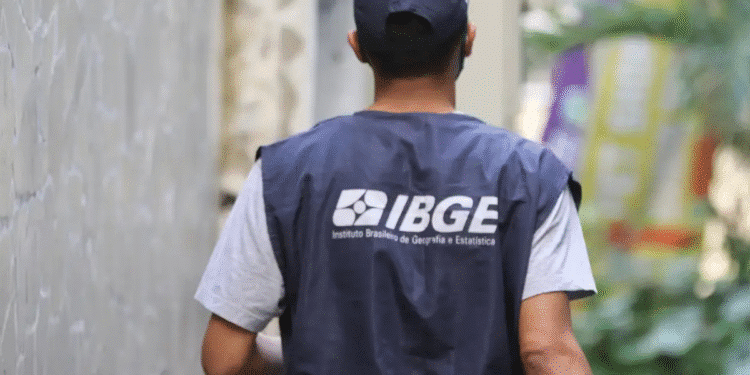 Agente do IBGE de costas com colete azul escuro e boné, caminhando entre prédios para coleta de dados