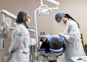 Duas dentistas com avental branco e máscaras atendendo paciente em cadeira odontológica profissional