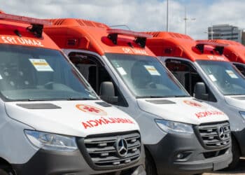 Várias ambulâncias do SAMU com pintura branca e vermelha alinhadas em fila sob céu azul com prédios ao fundo.