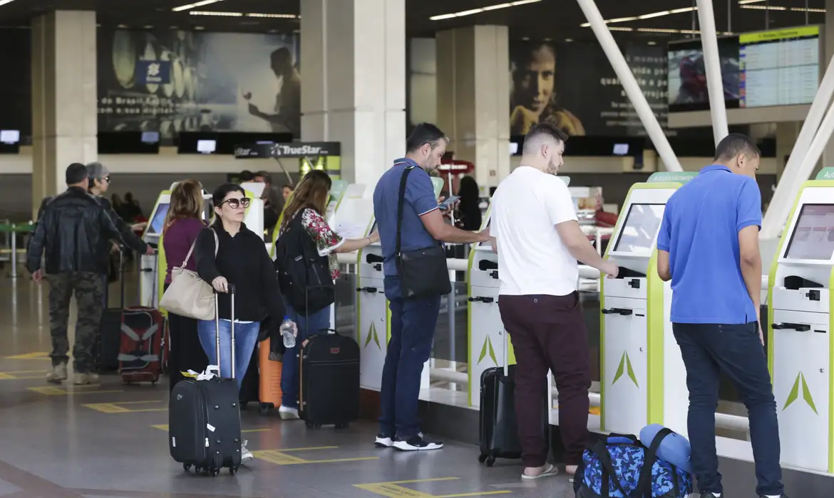 Pessoas em fila utilizando totens de autoatendimento para check-in no aeroporto com malas e mochilas