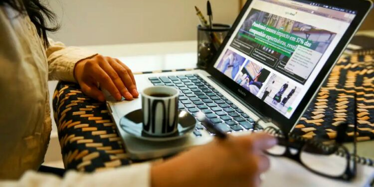 Mulher em casa em teletrabalho digitando no notebook e fazendo anotações em uma folha, com xícara de café ao lado, simbolizando a flexibilidade da carreira híbrida.