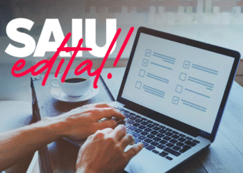 Imagem ilustrativa de mãos digitando em um laptop com tela exibindo checklist, xícara de café ao lado e texto overlay "SAIU edital!" em rosa e branco.