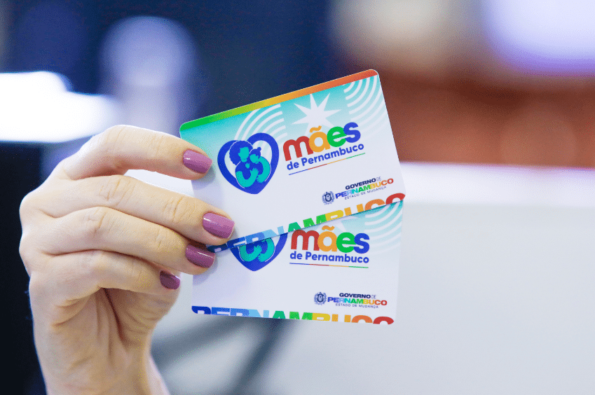 Auxílio de R$ 300 do programa Mães de Pernambuco Mão segurando cartões coloridos do programa Mães de Pernambuco, com logo e texto do Governo do Estado.