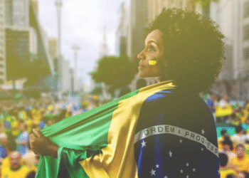 Mulher com rosto pintado, envolta na bandeira do Brasil em manifestação na rua.