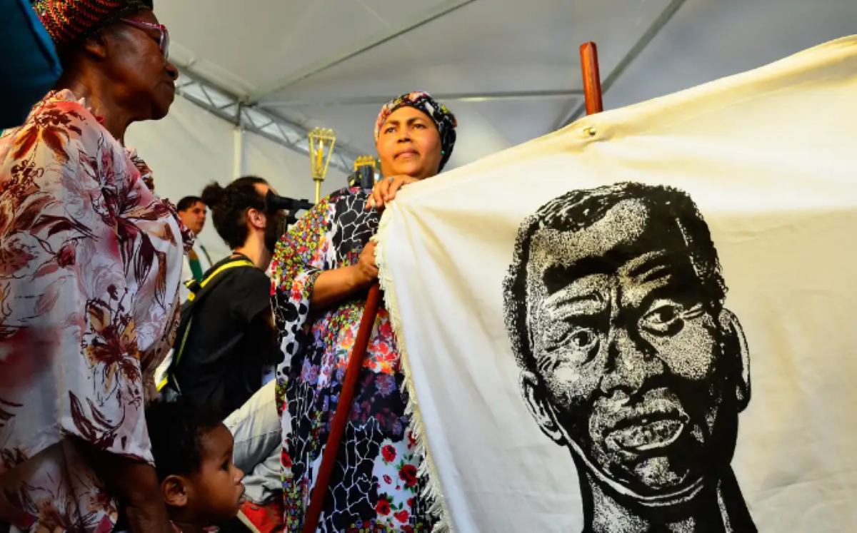 Mulheres negras seguram estandarte com retrato em preto e branco de homem negro, em evento sob tenda.