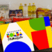 Cartão do Programa Bolsa Família em primeiro plano com fundo desfocado mostrando uma variedade de alimentos básicos (arroz, óleo, café, farinha), simbolizando o auxílio governamental a famílias de baixa renda.