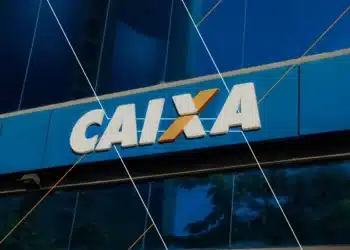 Letreiro da Caixa Econômica Federal branco e laranja em prédio com vidro azul refletindo árvores
