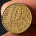Close-up de uma moeda brasileira de 10 centavos do ano 2010 sendo segurada, destacando o defeito de fabricação conhecido como cunho quebrado (a trinca na superfície). A imagem ilustra a raridade numismática que pode ser negociada por R$ 60 ou mais.
