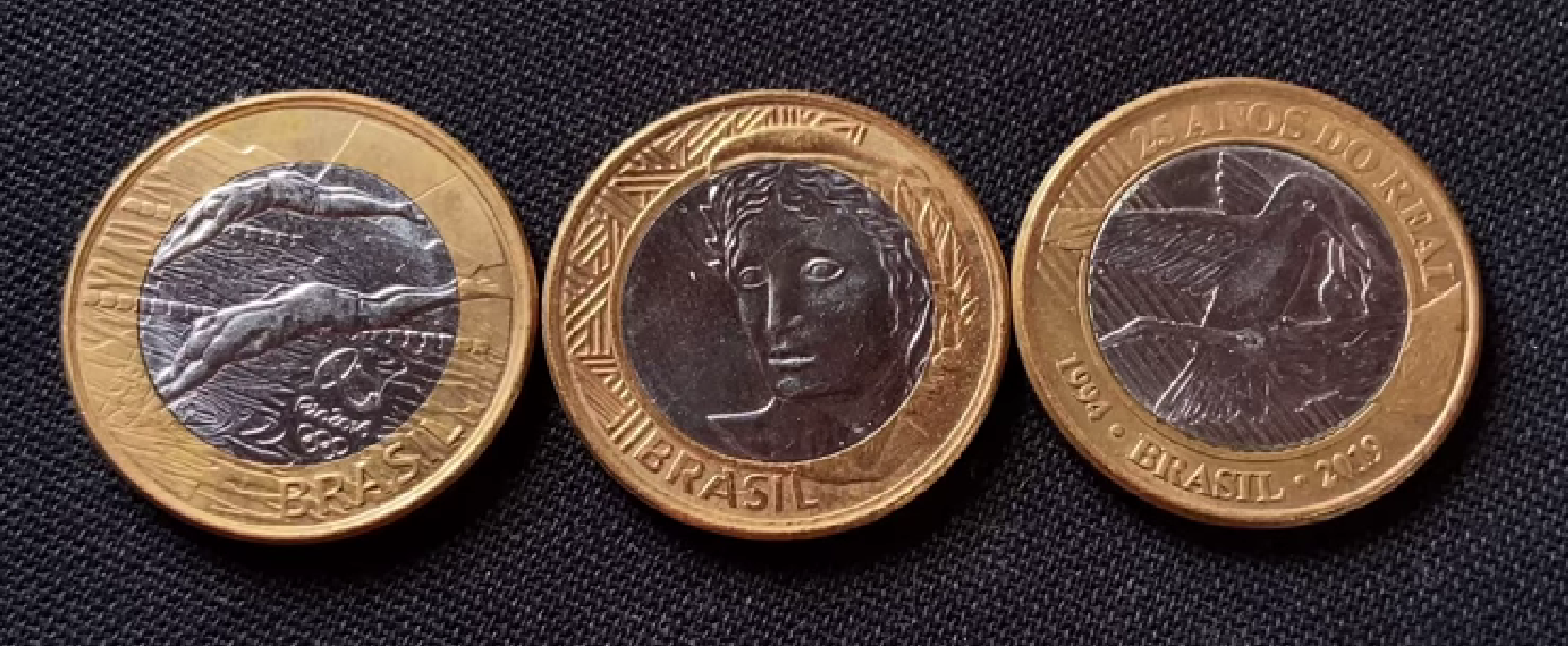 Três moedas brasileiras de 1 Real: uma comemorativa (25 Anos do Real), uma comum (Efígie da República) e uma da série Olimpíadas (Natação), sobre fundo escuro.