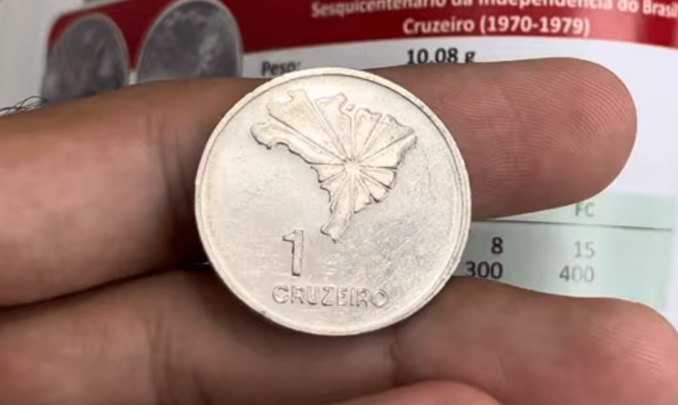 Moeda de 1 Cruzeiro de 1972: Raridade do Sesquicentário Moeda brasileira de 1 Cruzeiro, datada de 1972, com o mapa do Brasil no anverso, sendo segurada. No fundo, parte de um catálogo numismático. Ilustra a moeda comemorativa do Sesquicentário da Independência que pode valer até R$ 700 devido a variações de erro, como bordo liso ou acabamento espelhado.