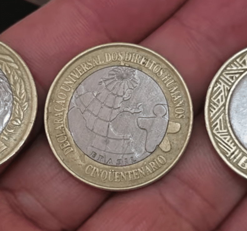 Moeda comemorativa de 1 Real de 1998, alusiva aos Direitos Humanos (DH), sendo exibida na palma da mão, reconhecida por sua tiragem limitada e alto valor de colecionismo.