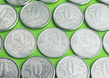 Close-up de diversas moedas de 50 centavos de diferentes anos (incluindo 1994, 2002 e 2016) sobre um fundo verde, representando a procura por anomalias e moedas raras no troco do dia a dia.
