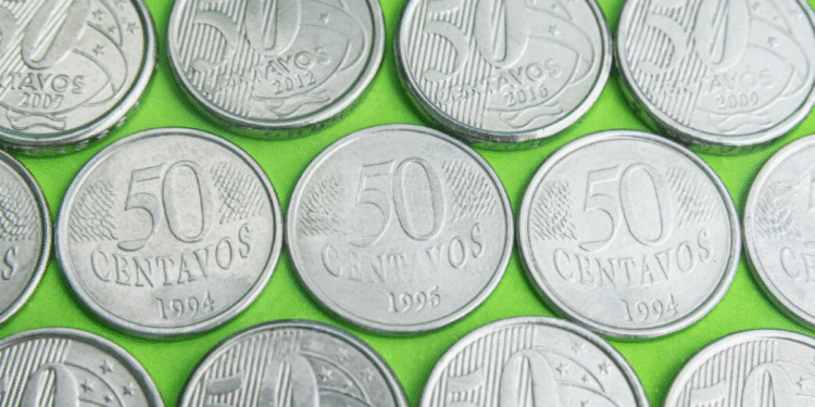 Close-up de diversas moedas de 50 centavos de diferentes anos (incluindo 1994, 2002 e 2016) sobre um fundo verde, representando a procura por anomalias e moedas raras no troco do dia a dia.
