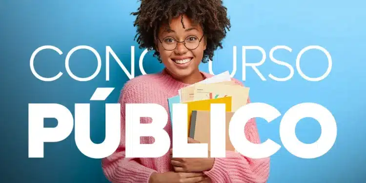 Jovem sorridente com óculos e suéter rosa segurando livros e papéis para concurso público.