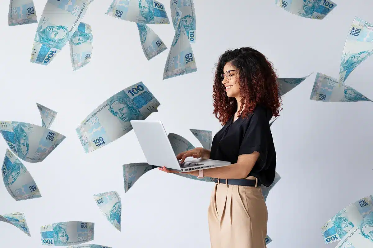 Mulher sorridente de cabelo encaracolado usando óculos e segurando notebook cercada por cédulas de 100 reais voando