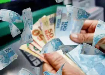 Notas de reais sendo contadas em frente a um caixa eletrônico, simbolizando benefícios e pagamentos de fim de ano.