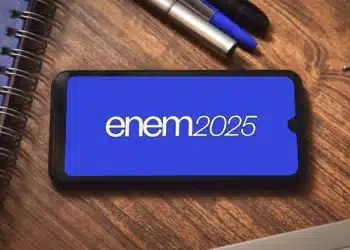 Celular exibindo a inscrição "enem 2025" em tela azul, apoiado sobre uma mesa de madeira com cadernos e canetas ao lado, simbolizando o exame e seus preparativos.