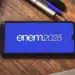 Celular exibindo a inscrição "enem 2025" em tela azul, apoiado sobre uma mesa de madeira com cadernos e canetas ao lado, simbolizando o exame e seus preparativos.