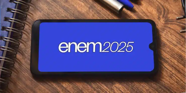 Smartphone exibindo na tela azul a inscrição "enem 2025" em branco, sobre uma mesa de madeira com cadernos e canetas.