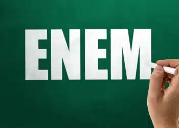 Mão segurando giz escrevendo a sigla ENEM em letras grandes e brancas sobre um quadro-negro verde-escuro. Exame Nacional do Ensino Médio.