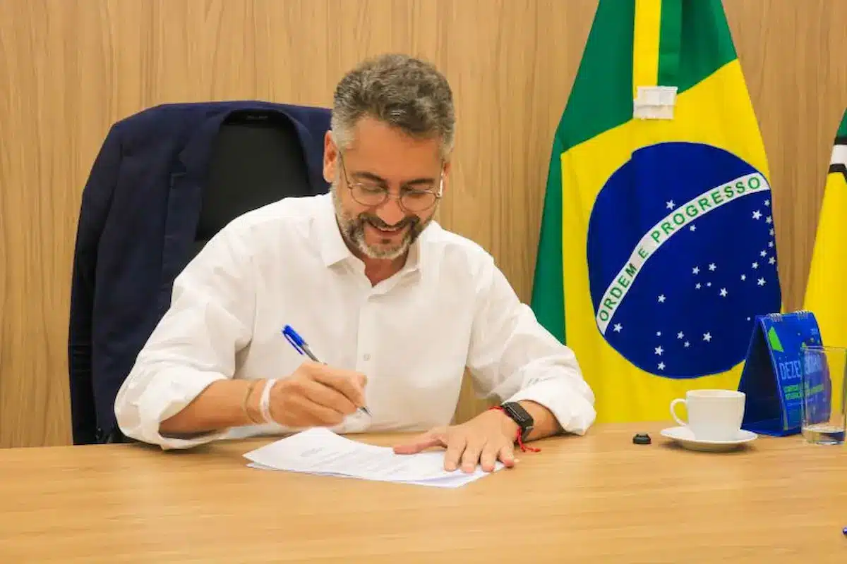 Governador do Amapá assinando documentos em gabinete, com bandeira do Brasil ao fundo.
