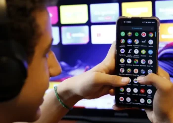 Pessoa segura smartphone mostrando tela de aplicativos enquanto usa fone de ouvido perto de TV
