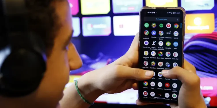 Pessoa segura smartphone mostrando tela de aplicativos enquanto usa fone de ouvido perto de TV
