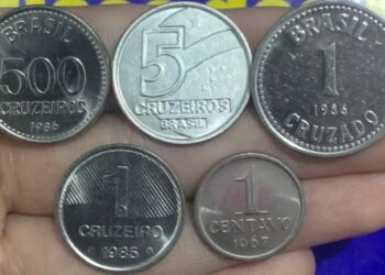 Cinco moedas antigas brasileiras em uma mão, incluindo exemplares de 500 Cruzeiros, 5 Cruzeiros, 1 Cruzado, 1 Cruzeiro e 1 Centavo de 1967. Ilustra a raridade das moedas de Cruzeiro e Cruzado que podem valer até R$ 2.000 devido a defeitos ou baixa tiragem.