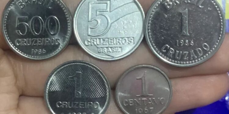 Cinco moedas antigas brasileiras em uma mão, incluindo exemplares de 500 Cruzeiros, 5 Cruzeiros, 1 Cruzado, 1 Cruzeiro e 1 Centavo de 1967. Ilustra a raridade das moedas de Cruzeiro e Cruzado que podem valer até R$ 2.000 devido a defeitos ou baixa tiragem.
