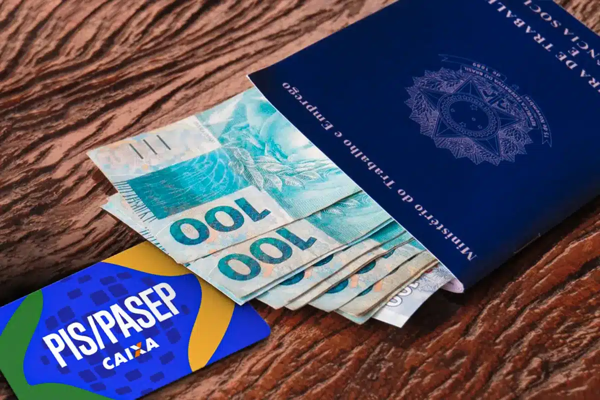 Carteira de trabalho azul, dinheiro em cédulas de cem reais e cartão PIS/PASEP sobre mesa de madeira
