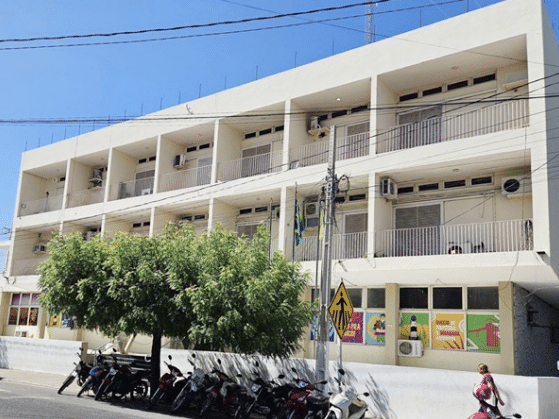Fachada do prédio da Prefeitura de Caicó, RN, com árvore, motos estacionadas, bandeira brasileira e murais coloridos na parte inferior.