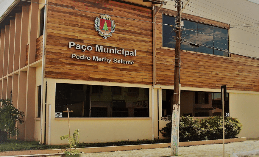 Concurso Prefeitura de Três Barras-SC Foto da fachada do Paço Municipal Pedro Mery Seleme em Três Barras, SC, com parede de madeira, emblema municipal e janelas envidraçadas, sob céu parcialmente nublado.